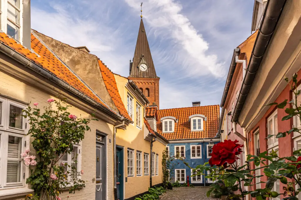 Charmante, farbenfrohe Häuser säumen eine gepflasterte Gasse in Aalborg, Dänemark, mit einer Kirchturmuhr im Hintergrund.